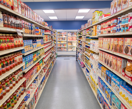 Wide grocery store aisle with fully stocked shelves on both sides, lined with boxed and canned foods, leading toward a freezer case at the back of the store under bright overhead lighting.