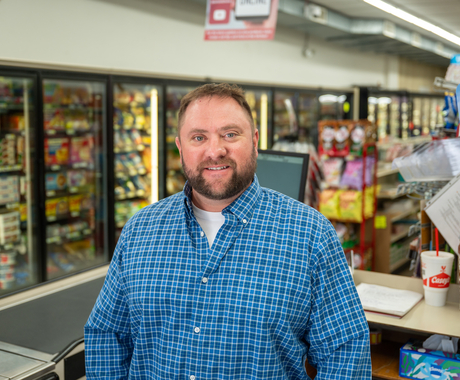 Man standing in a grocery store at a check-out stand. In the background are coolers with frozen food. Man has facial hair, short hair, and is wearing a long-sleeved blue plaid shirt.