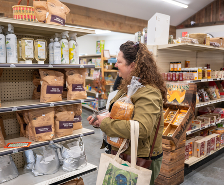 Woman with curly hair, wearing a coat, holds a loaf of bread and a tote bag. She's in a grocery store looking at a shelf of flour and oils.