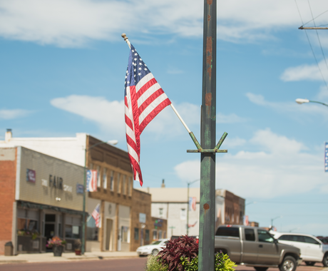 An American flag waves, suspended from a weathered green post on a rural main street. In the background, we see vehicles parked on a brick road and historic brick-front buildings. There is a potted plant below the flag and the sky is bright blue with wispy clouds.