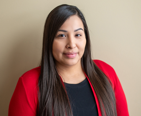 Native American woman with long black hair past her shoulders, looks at the camera, wearing a long sleeve red cardigan with a black shirt underneath, standing in front of a beige wall
