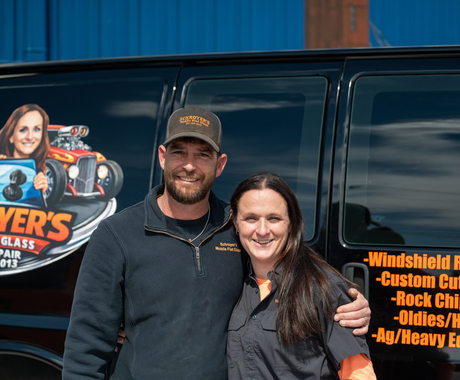 A woman and a man stand in front of a black van. On the van is an image that says Schroyer's Mobile Flat Glass, windshield replacement, custom cut flat glass, rock chip repair, oldies/hot rods, ag/heavy eq. The man is wearing a dark colored fleece with a collar and a ball cap. The woman has long straight brown hair and is wearing a button up short sleeve shirt.