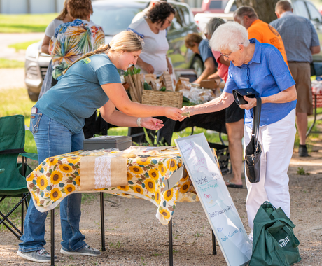 A young women with a long blonde braid wearing a blue t-shirt and jeans accepts money from an elderly women with white hear wearing a blue button-down short-sleeved shirt with white pants. It's a sunny day at the farmers market and there are a handful of other shoppers and vendors in the background. The young woman and elderly women are exchanging money over a folding card table, covered in a sunflower table cloth.