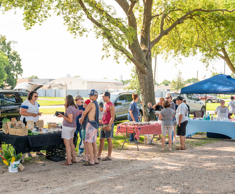 A row of farmers market vendors along a road. Vendors have tables with bright colored clothes and products on top. Groups of customers are in front of each table.
