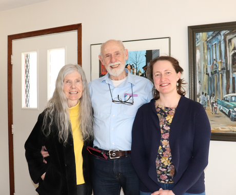 Three people standing side by side indoors, smiling at the camera. On the left is an older woman with long gray hair, wearing a yellow turtleneck and black jacket. In the center is an older man with a white beard, dressed in a light blue button‑down shirt with glasses hanging from his collar. On the right is a younger woman with brown hair pulled back, wearing a navy cardigan over a floral blouse. Behind them is a white wall with framed artwork and a doorway.