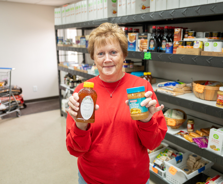 White female stands in front of food pantry shelves holding a jar of honey and jar of peanut butter