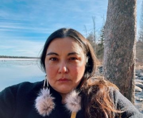 Portrait from the shoulders up of a woman with long dark hair wearing a black coat and fluffy pom‑pom earrings, standing beside a tree on a calm lakeshore under a clear blue sky.
