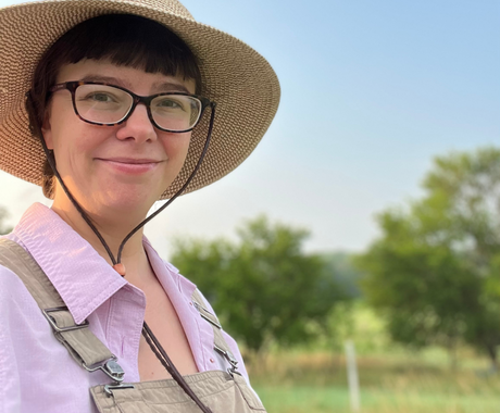Woman with short dark hair, a sunhat, and dark glasses, looking at the camera. She is also wearing a collared pink shirt and beige overalls. In the background is a blue sky and green trees and green grass.