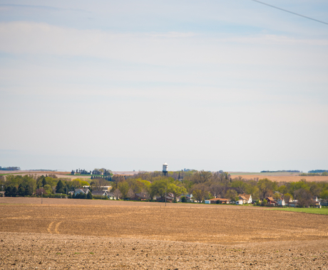 Small town with a handful of bright green trees in the springtime. In the foreground is a field ready to be planted. In the middle of town is a water tower. You can see more fields on the other side of the town in the background.