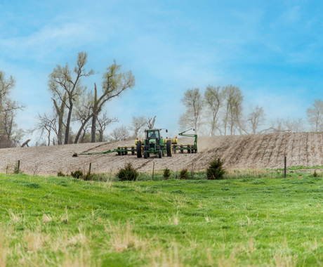 Fotografía de un tractor que se dirige hacia nosotros en un campo sin labranza. En primer plano se ve hierba verde. El tractor es un John Deere verde con una sembradora ancha detrás. Al fondo se ven árboles con brotes y hojas pequeñas.