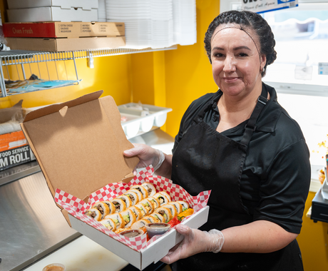 A dark-haired woman wearing a black hair net, black t--shirt, and black apron smiles at the camera while holding a cardboard box of Mexican sushi.