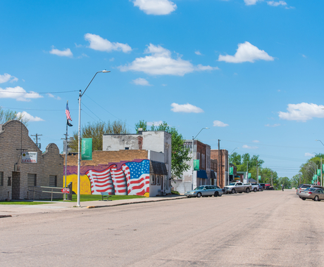 Rural Main Street, with a big flag mural on the side of a building on the left side of the street. About two blocks of downtown buildings with cars parked on both sides