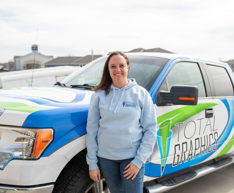 Una mujer blanca con cabello castaño hasta los hombros y vestida con una sudadera con capucha azul claro se para frente a un camión con diseños azules, blancos y verdes y las palabras "Total Graphics" en el lado del conductor.
