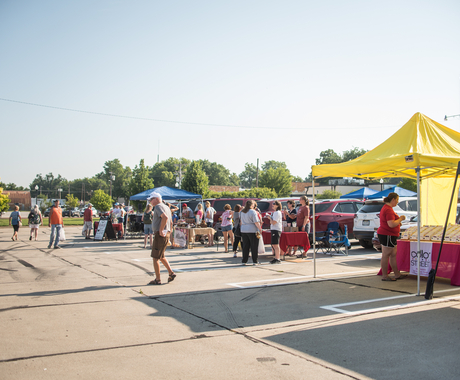 A crowd of people gathered in a parking lot with tents for a farmers market.