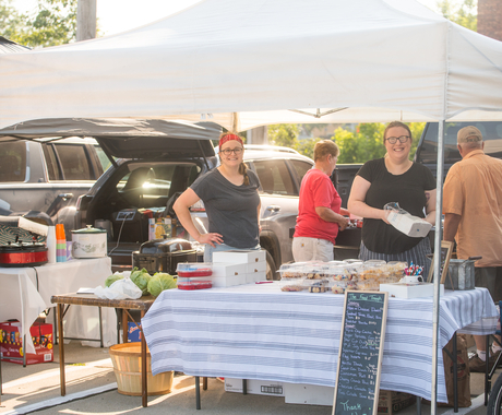 Two women standing at a table with baked goods with a tent over them at a farmers market