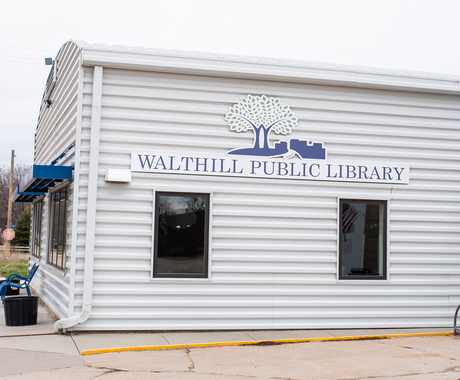 A large building with horizontal white metal siding. There are two windows with a blue sign above that says "WALTHILL PUBLIC LIBRARY". The sign also includes a silhouette of trees and a cityscape. Around the corner of the building, we see two blue awnings overhanging two sets of windows and a blue metal bench with black planters on both sides of it.