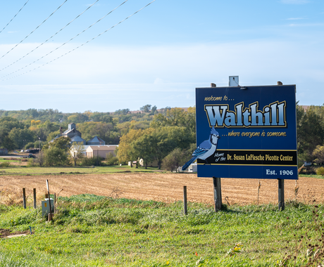 A roadside welcome sign for Walthill, Nebraska, stands beside a grassy field under a clear blue sky. The sign reads “Welcome to Walthill… where everyone is someone,” and notes it is the home of the Dr. Susan LaFlesche Picotte Center and established in 1906. Behind the sign are harvested fields, scattered trees, and a few buildings in the distance.