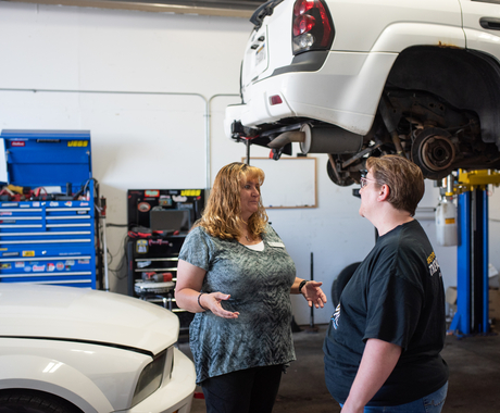 Two women standing in a car repair shop, with one car lifted with its wheels off