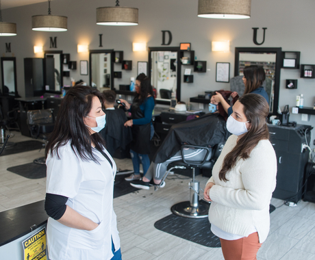 Two women with masks face to face at a beauty salon, with two women doing people's hair behind them