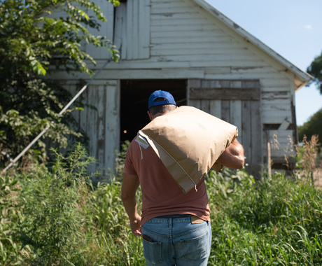 Man carrying a sack of feed with grass in the foreground and barn in the distance.
