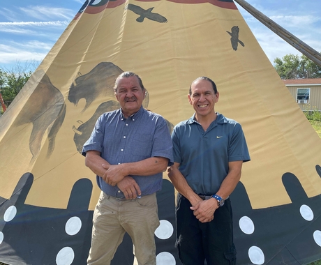 two Native American men wearing blue polos standing in front of a beige teepee with buffalo and birds on it