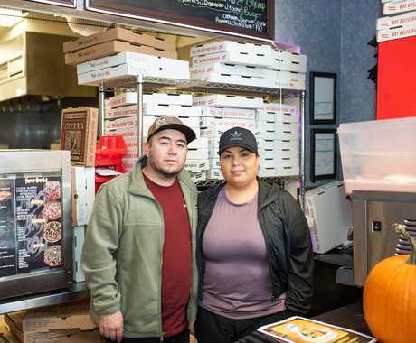 A man and a woman stand in front of a pile of pizza boxes behind them in a pizza shop