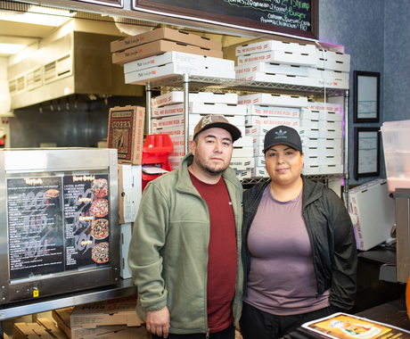 Un hombre y una mujer posan para una foto en frente de una repisa con cajas de pizza
