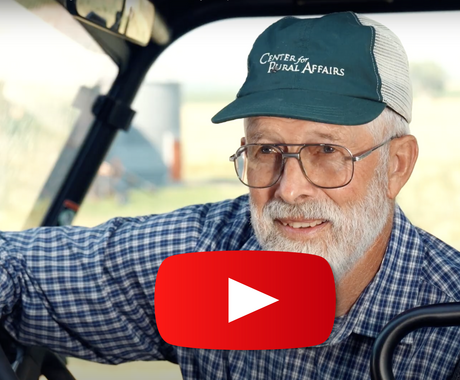 Smiling man with white mustache and beard leaning over a steering wheel of a side-by-side, wearing a blue plaid long-sleeved button up shirt and a green and white "Center for Rural Affairs" hat