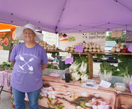 Woman in t-shirt that says "Little Town Gardens" in front of a vegetable stand  with bok choy, onions, and more