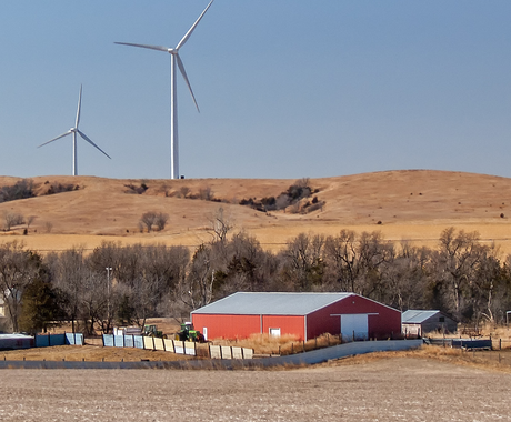 A rural landscape featuring a red barn with a gray roof surrounded by leafless trees and fields in late fall or winter. In the background, two large wind turbines stand on rolling, golden-brown hills beneath a clear blue sky. A green tractor is visible near the barn, and a fenced area borders the property.