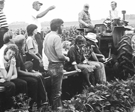 Fotografía histórica en blanco y negro de un grupo de agricultores y miembros de la comunidad reunidos en un campo de cultivo, sentados y de pie sobre equipos agrícolas y un tractor, mientras un hombre hace un gesto como si hablara a la multitud.