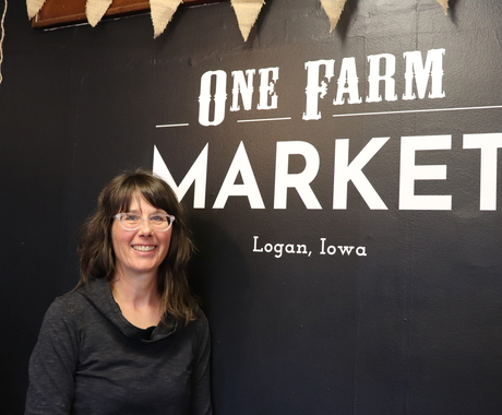 Woman in black shirt with clear glasses stands in front of black wall with white writing on it that says One Farm Market Logan, Iowa