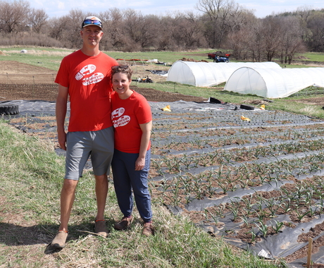 Man and woman in red t-shirts standing in a planted field with white tent greenhouses in background.