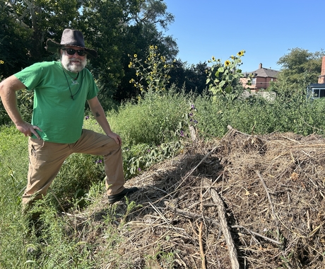 White bearded man wearing a green shirt, tan pants, a grey hat, and sunglasses stands on top of a mound of wood debris in a field