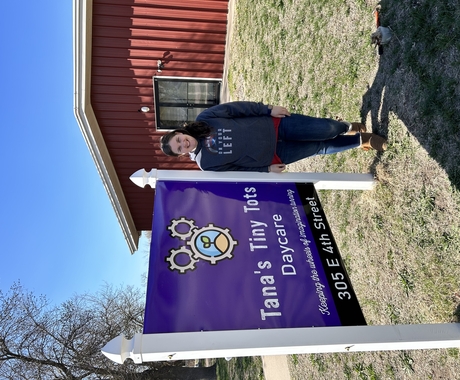 Woman in grey sweatshirt and blue jeans stands next to a purple business sign in front of a red building