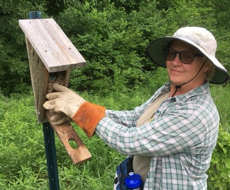 Woman in garden hat and heavy gloves, standing in front of a bird house box open containing a nest and eggs