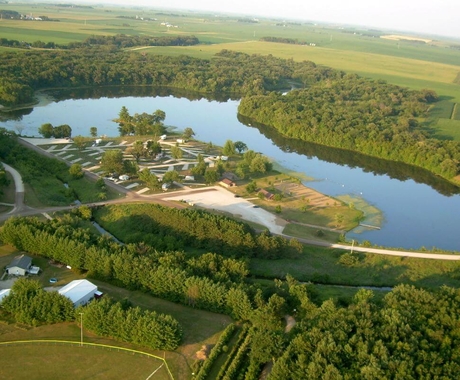 An aerial shot of a lake surrounded by green trees, then beyond that, green fields. On the left side of the lake is a campground with white roads and white camper pads, with a handful of travel trailers parked in spots.