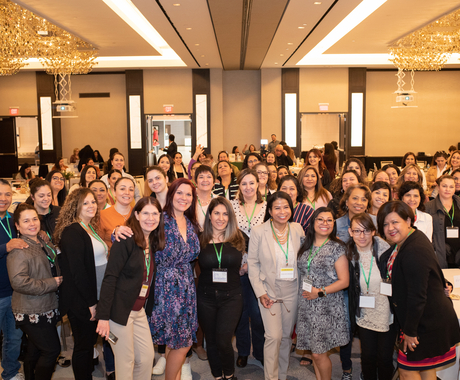 About 50 women dressed in business attire smiling at the camera in a hotel ballroom