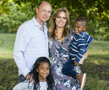 Family photo of a male, female, and two kids. Male and female are standing. Female is holding one child. The other child is standing in front of male. In a park with trees.