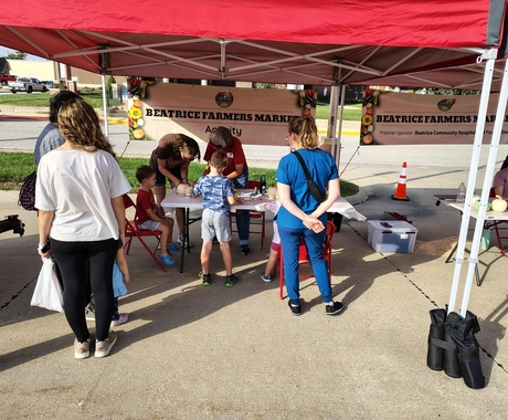 Several adults and several children gathered under a red tent. A sign behind the people reads, "Beatrice Farmers Market"