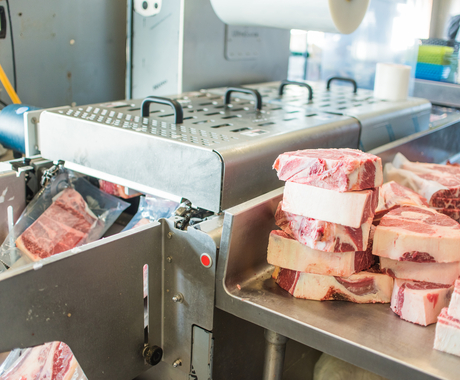 Meat processing photo - packaged meat coming out of plastic wrap machine and raw meat stacked on counter