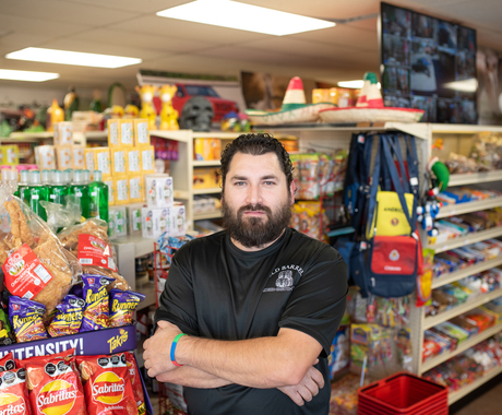 Hombre con barba y camisa negra posa para una foto con brazos cruzados en frente de mercancía en una tienda