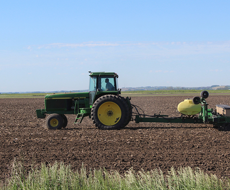A green John Deere tractor heads across a freshly plowed field with a planter attached to the back. A small plume of dust is behind the trailer.
