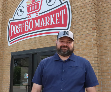 A man in a navy blue button down and a grey Nebraska baseball cap smiles at the camera outside of a brown brick building with a red and white sign that reads "Post 60 Market."