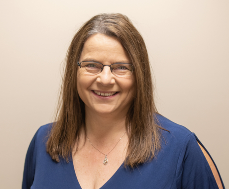 Woman with brown hair and glasses smiling at the camera, wearing a blue shirt and a necklace