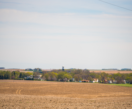 Landscape photo of a harvested field in foreground with small town with fall trees in the middle of the photo. A water tower is in the very center, with its top above the trees.