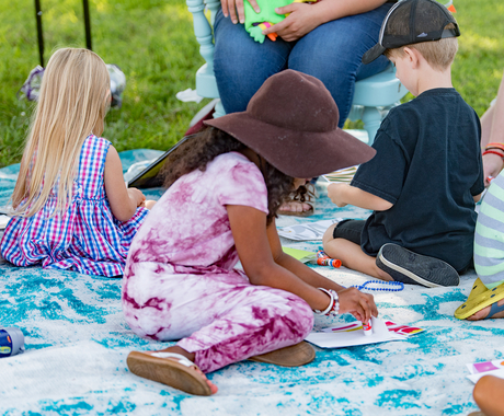 Children doing art projects while sitting on a blanket in a grassy area