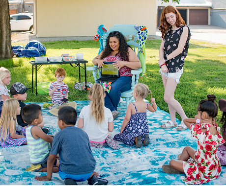 woman sitting in chair, book in hand, with another woman standing, both looking at seated children