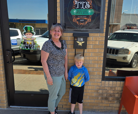 A woman and a young boy stand outside a brick building beneath a sign that reads “TTS Hometown Pub & Grub, Established 2024.” The woman is smiling and wearing a patterned black and white shirt with light green pants, while the boy wears a blue hoodie with a yellow flame design and black shorts. The entrance door next to them has a decal featuring a child in a leprechaun hat and the same business name. 