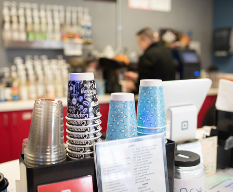 A row of cups, clear, white and black and blue with white are at the front while a man works a coffee machine in background